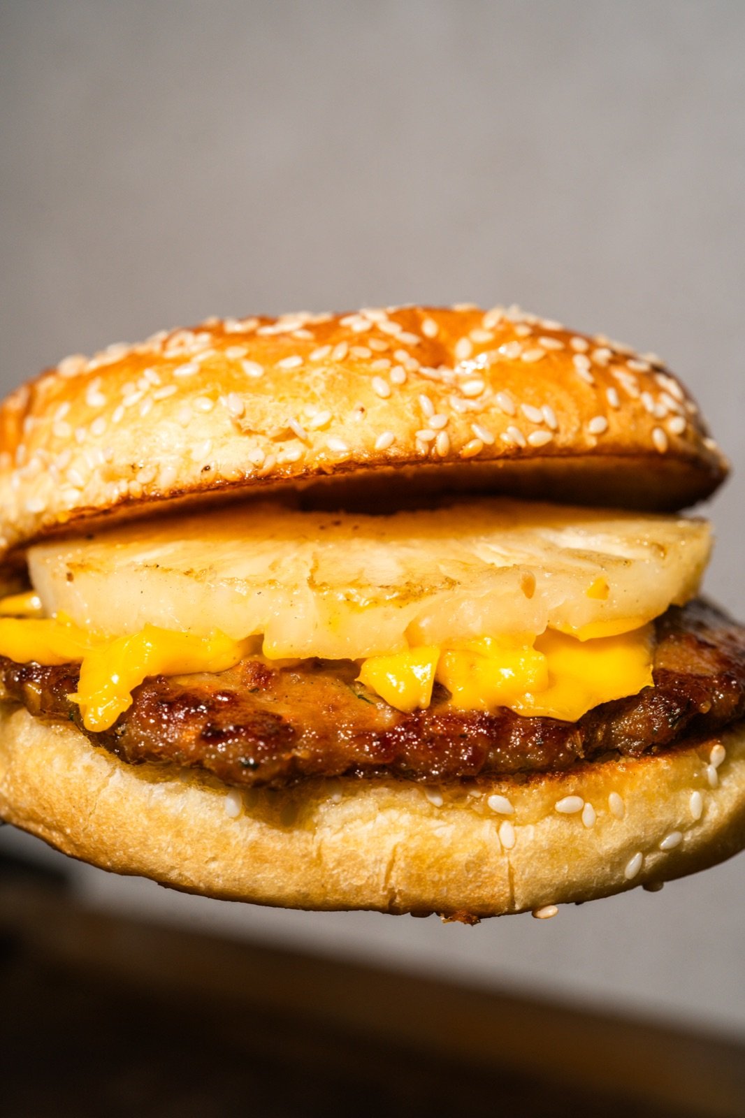 Burger completa con piña dorada y queso escurriendo, sobre fondo gris neutro